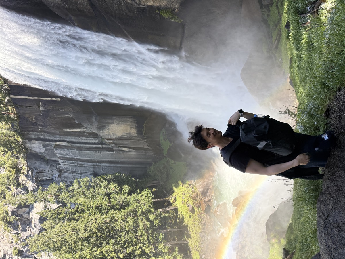 David C. Jeong smiling in front of Vernal Fall in Yosemite National Park, with a rainbow in the mist.