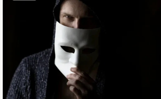 A person in a hood holding a plain white mask in front of their face.