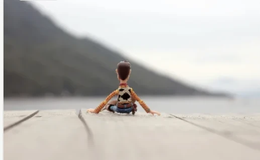 A small toy figure sitting alone at the end of a wooden dock, looking out at the water.